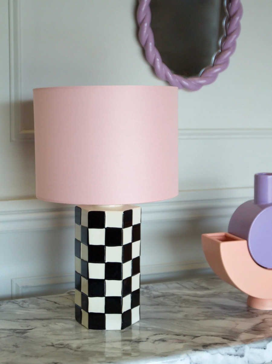 Lámpara de estilo kitsch, con base de patrón de ajedrez o damero en blanco y negro, y pantalla rosada. Mesa de mármol en tonos grises. Espejo moderno con marco color lila.