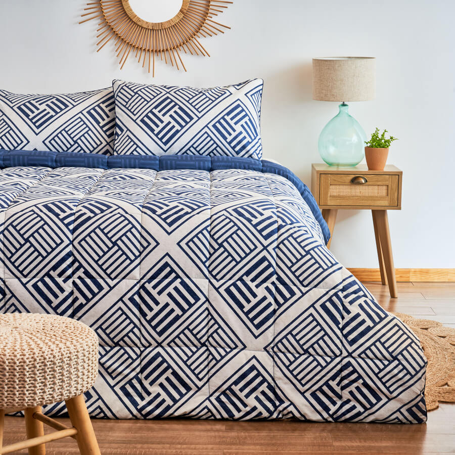Cama king en dormitorio moderno. Plumón geométrico en colores blanco y azul. Piso de madera con base tejida, alfombra redonda de yute y velador de madera.