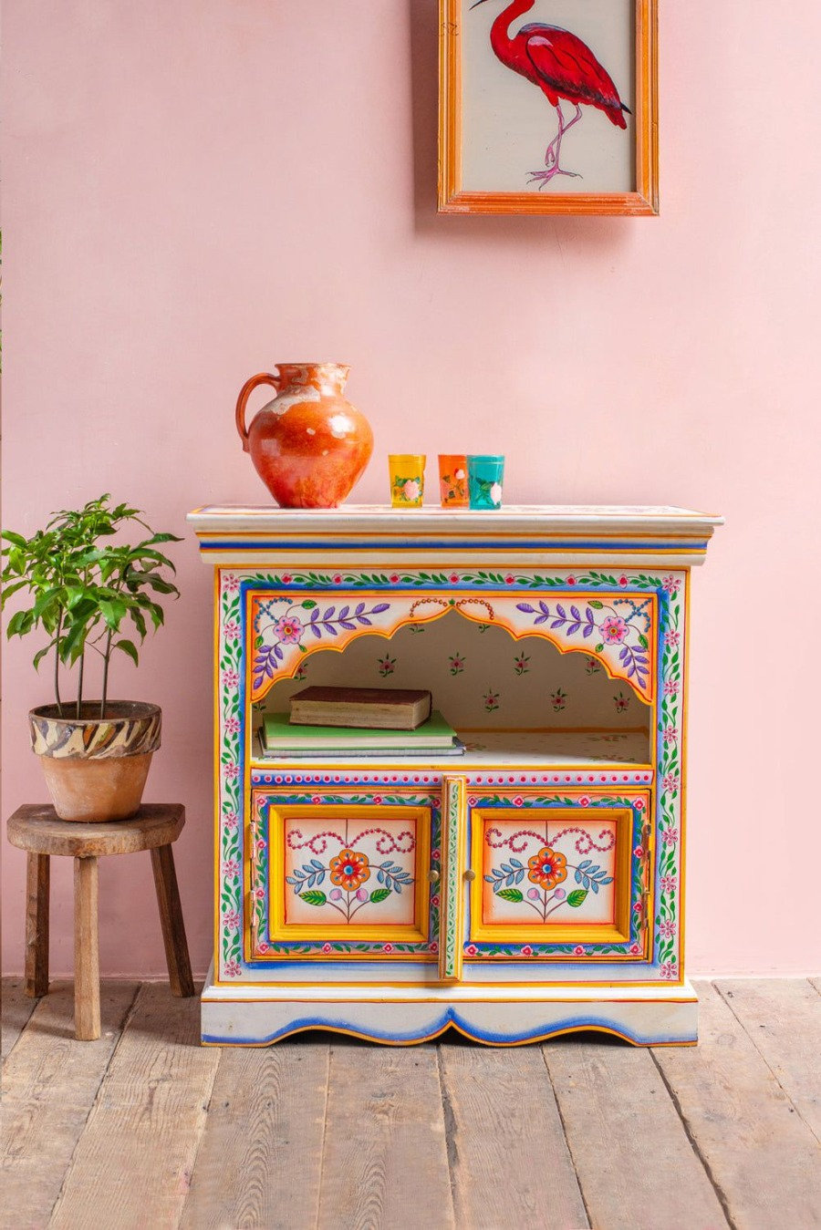 Mueble vintage blanco pintado con motivos de flores y distintos patrones. Sobre el mueble hay un jarrón naranjo y tres vasos.