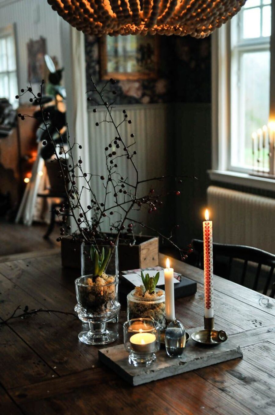 Detalle de mesa estilo rústico. Mesa de madera, con velas decorativas, plantas en maceteros de vidrio y una caja de madera. Ramas secas. Gran lámpara de techo con pelotas de madera.