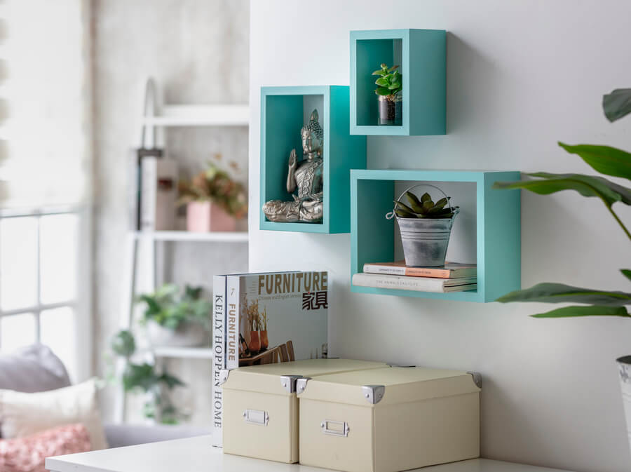 Detalle de una pared de un escritorio. 3 repisas flotantes, tipo cubo, de color turquesa. Dos cajas organizadoras de color beige, revistas, adornos y dos plantas suculentas.