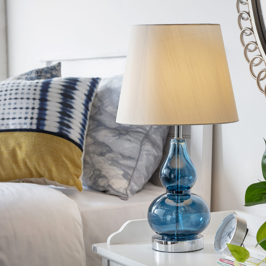 Lámpara de mesa con pantalla color crema y base de vidrio azul. Está sobre un velador blanco al lado de una cama.