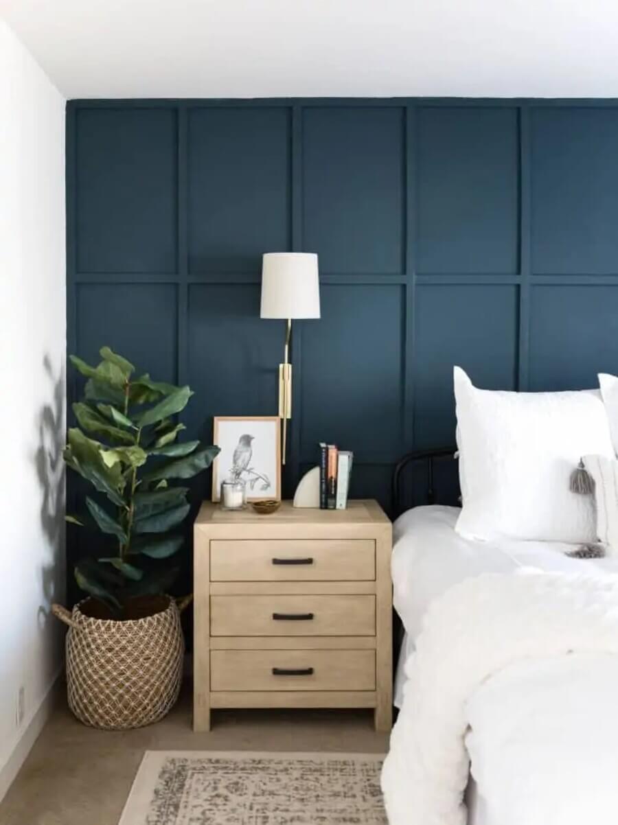 Dormitorio moderno, con pared forrada con paneles de madera, de color azul. Velador de madera con tres cajones, junto a él una planta en un canasto. Cama con sábanas y plumón blanco. Lámpara de muro dorada con pantalla de tela color blanco.