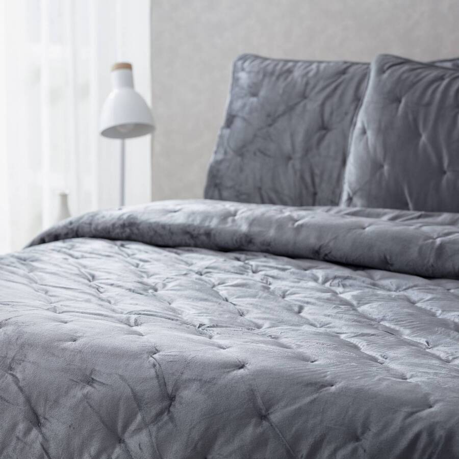 Detalle de cama de dos plazas con plumón acolchado aterciopelado color gris. Dos almohadas con fundas de la misma tela. Al fondo se ve una lámpara de mesa blanca de metal estilo nórdico