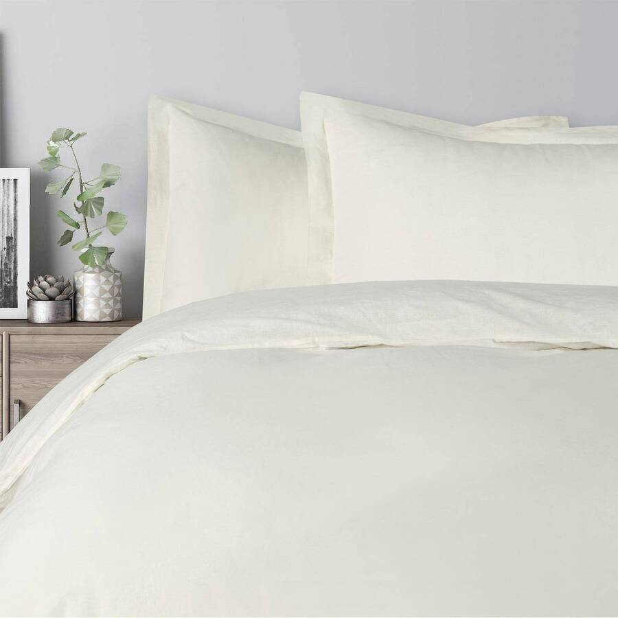 Cama queen con plumón color crema, dos almohadas también de color crema. Pared gris claro, velador de melamina con maceteros metálicos con plantas