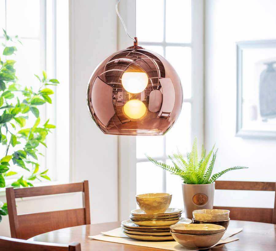 Gran lámpara con pantalla redonda de vidrio color café, estilo mid century. Mesa redonda de madera con tres sillas de madera, Vajilla de cerámica y planta en maceta pequeña. 