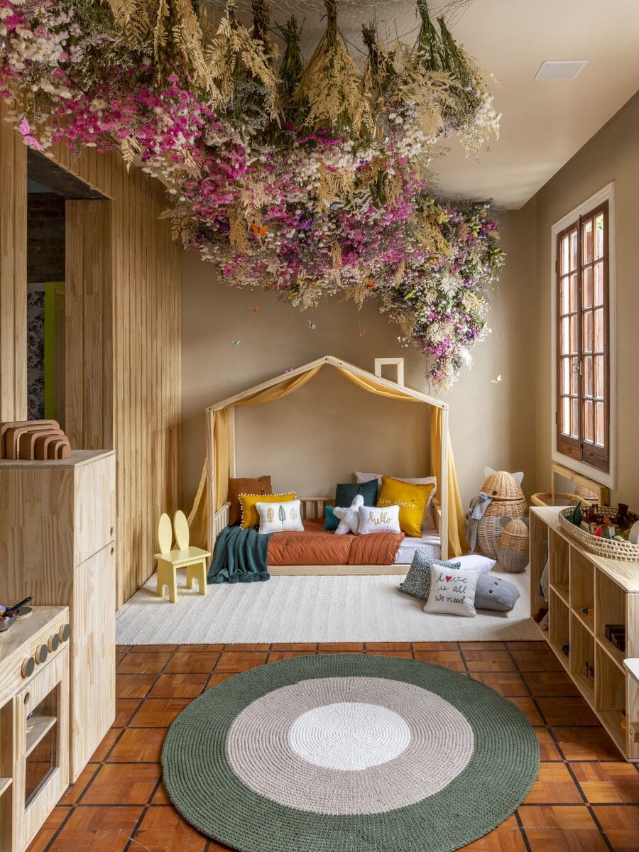 Dormitorio Montessori con una cama con marco de madera en forma de casita, una alfombra blanca y otra circular, varios estantes y juguetes de madera. El techo está cubierto con fñores secas.