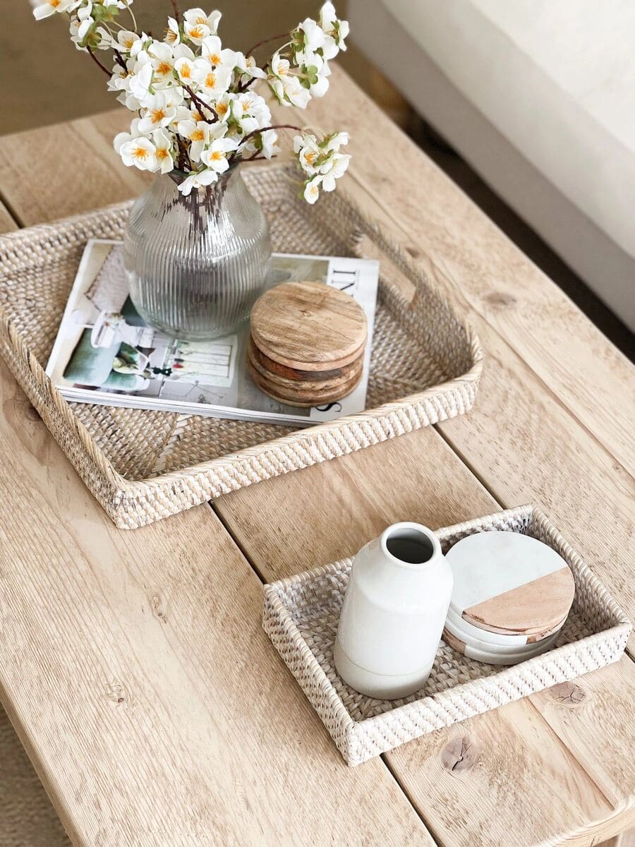 Dos bandejas de fibras naturales de distintos tamaños en una mesa de centro. Las bandejas contienen jarrones con flores, portavasos y otros adornos.
