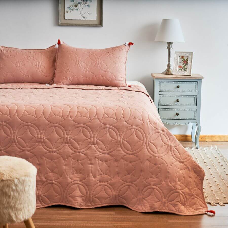 Cama queen, con cubrecama y almohadas de color rosa. Junto a la cama hay un velador estilo provenzal, de madera, color celeste claro y tres cajones. Hay una lámpara clásica de mesa, de base plateada y pantalla blanca. Junto a la cama hay una bajada de cama o alfombra pequeña de fibras naturales.