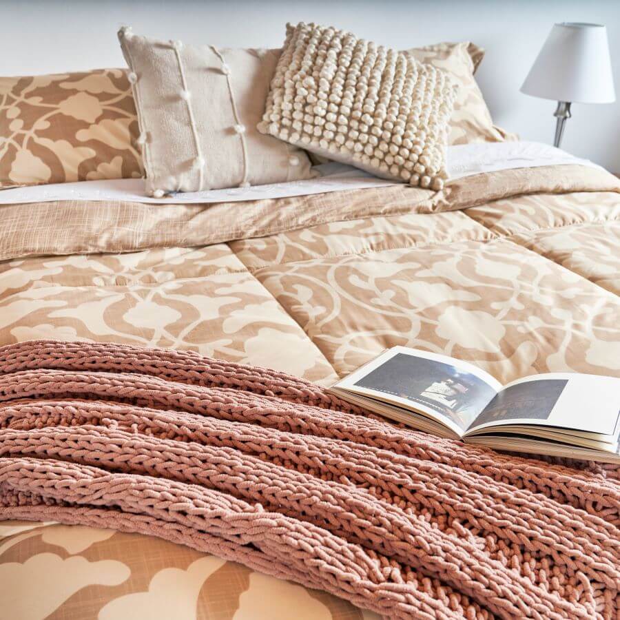 Detalle de una cama queen, con cubrecama con diseñi de flores, en tonos beige y mostaza. Tiene dos almohadas y dos cojines cuadrados. Los dos son de color beige y tienen detalles con pelotitas de lana. Piecera de color rosa.