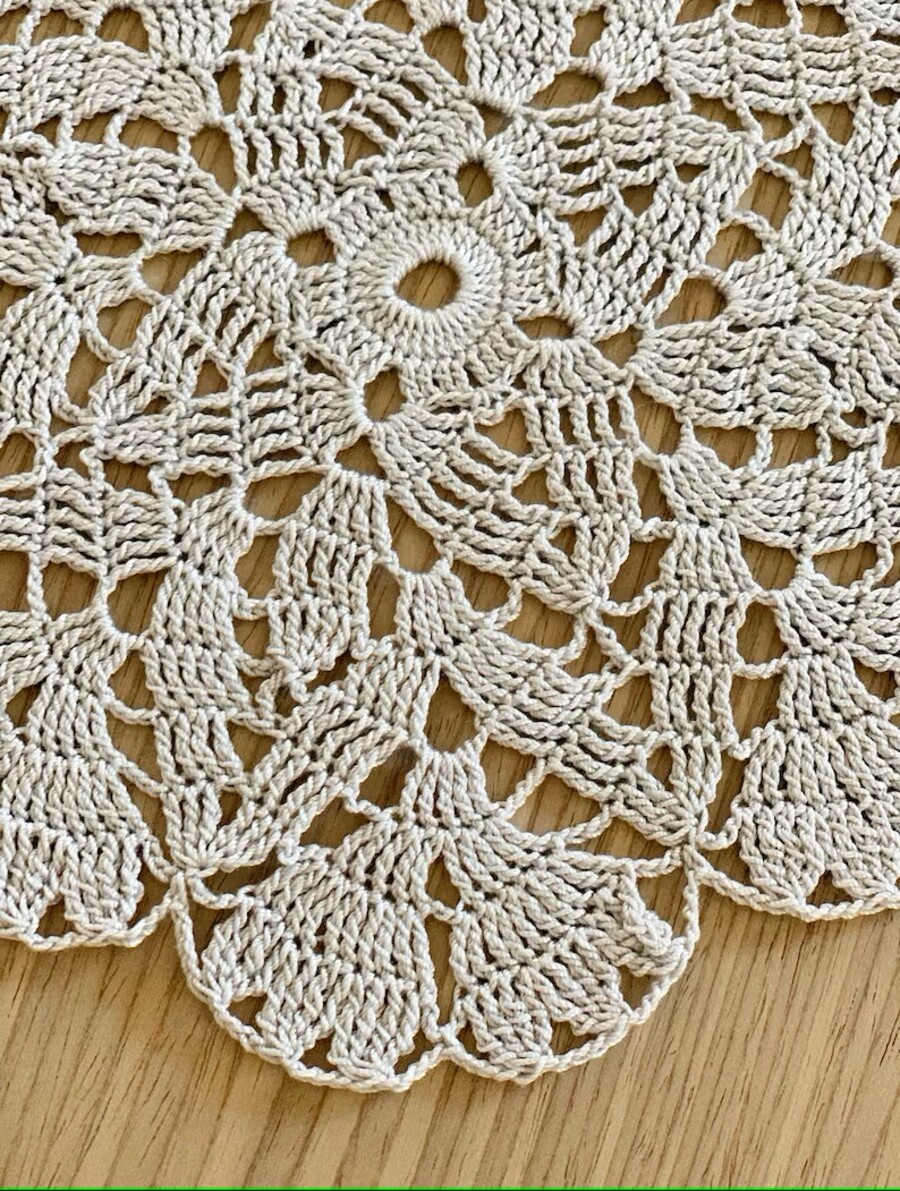 Detalle de un paño tejido a crochet, con forma de flor, hecho con hilo color beige o crudo. Está sobre una mesa de madera.