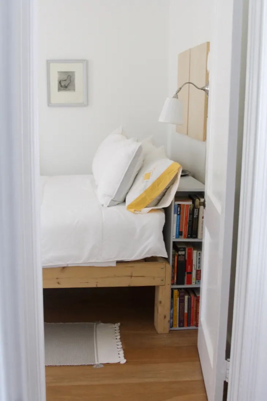 Cama de dos plazas con base de madera. Detrás de la cama hay un mueble angosto de madera con varios libros. Lámpara de muro metálica con pantalla blanca. Las paredes son blancas y el piso es de madera.