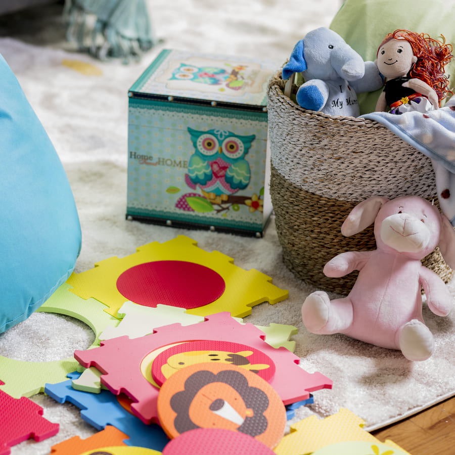 Alfombra blanca y, sobre ella, piezas de puzzle de goma eva con diseño de animales, juguetes y un canasto de fibra natural con más juguetes y una manta.