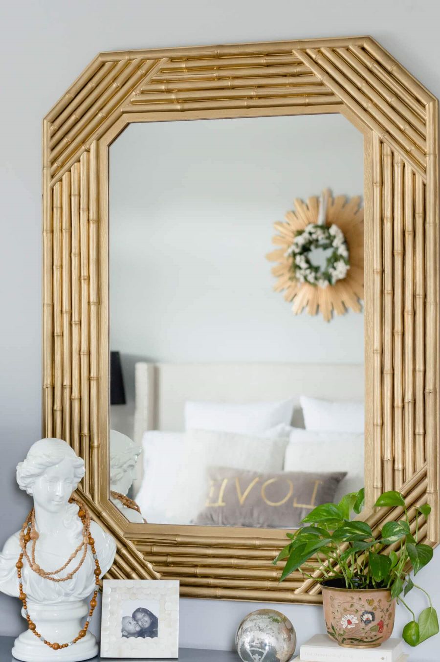 Espejo con marco de bambú, pintado de color dorado. Está sobre una repisa con un busto blanco, con un collar de madera, un pequeño marco de foto y un macetero pintado con flores. En el espejo se ve reflejada una cama blanca, con un cojín que dice "love".