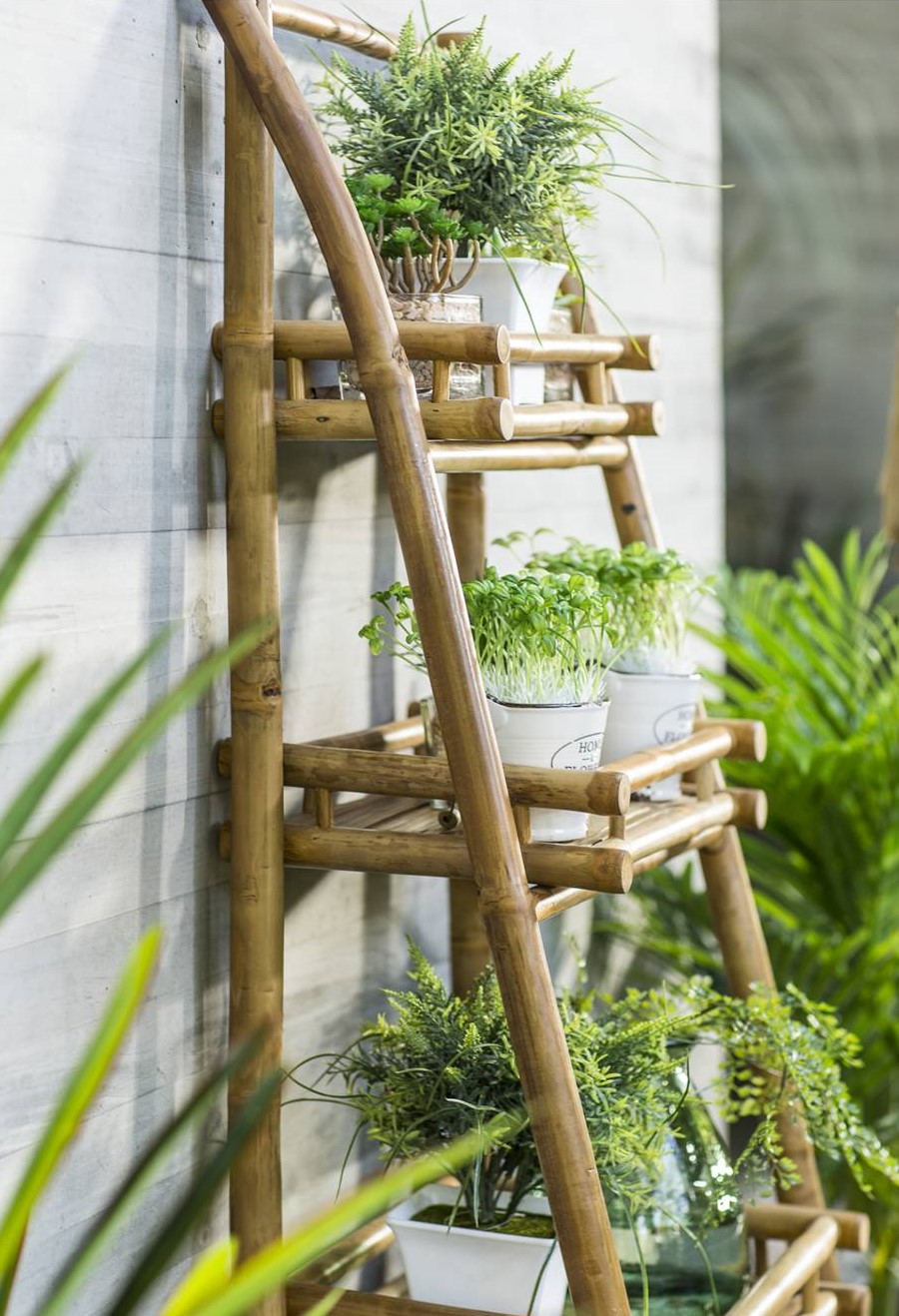 Detalle de una repisa de tres niveles, de bambú en una terraza. En ella hay varias macetas blancas con hierbas y plantas, La repisa está apoyada en un muro gris.