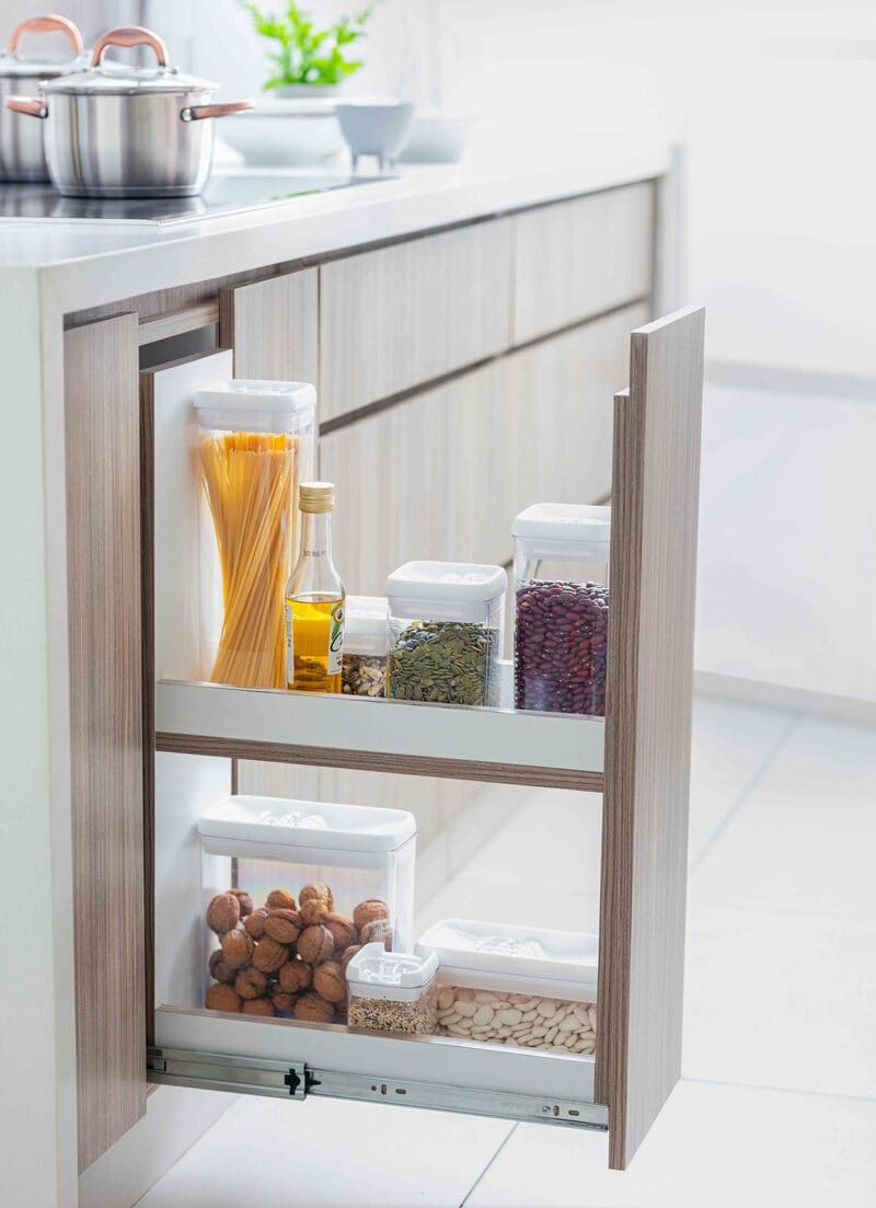 Mueble de cocina de madera y cubierta blanca. Hay un cajón angosto extraíble con contenedores transparentes con ingredientes.