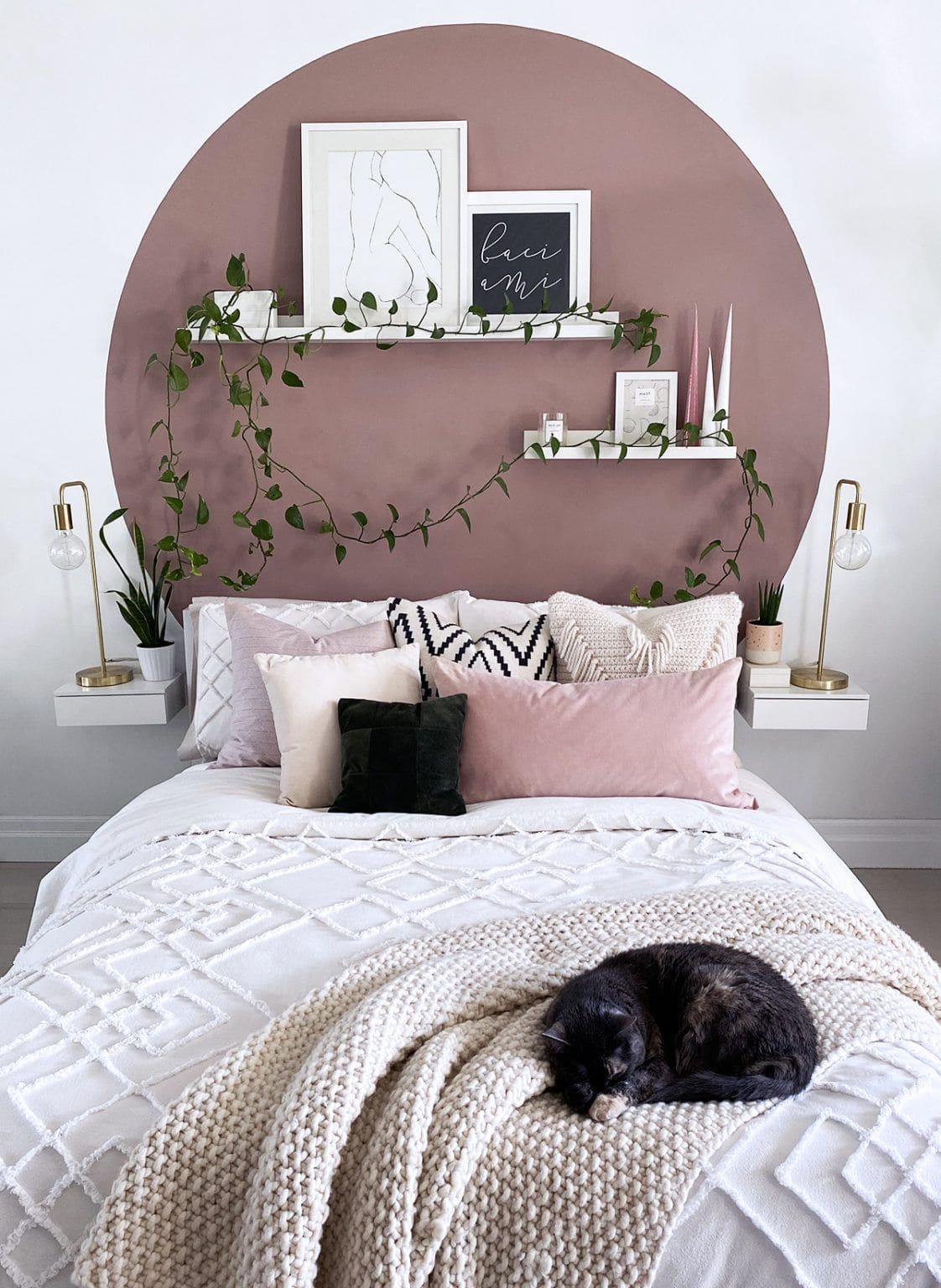 Dormitorio con una cama con frazada blanca y cojines. En su cabecera, hay un círculo color malva pintado en la pared y, a cada lado, una repisa blanca con una lámpara de mesa dorado. En el círculo también hay repisas blancas con cuadros y plantas.
