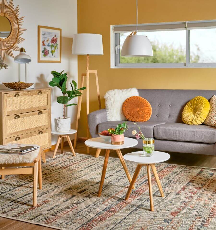 Living de estilo boho. Un muro color amarillo mostaza, y otro muro color blanco. Sofá de dos cuerpos, color gris, de estilo nórdico. Tiene 4 cojines, uno planco peludos, dos redondos de color amarillo y naranjo y otro estampado con flores. Set de dos mesas de centro redondas, con base blanca y pata de madera. Gran alfombra con patrones geométricos desgastados. Cajonera de madera y fibras naturales con tres cajones, sobre ella un bowl de mimbre y en el muro un espejo redondo de mimbre con forma de sol. En la esquina hay una lámpara de pie con base de madera y pantalla blanca. En el techo hay una lámpara colgante de pantalla blanca, redondeada.