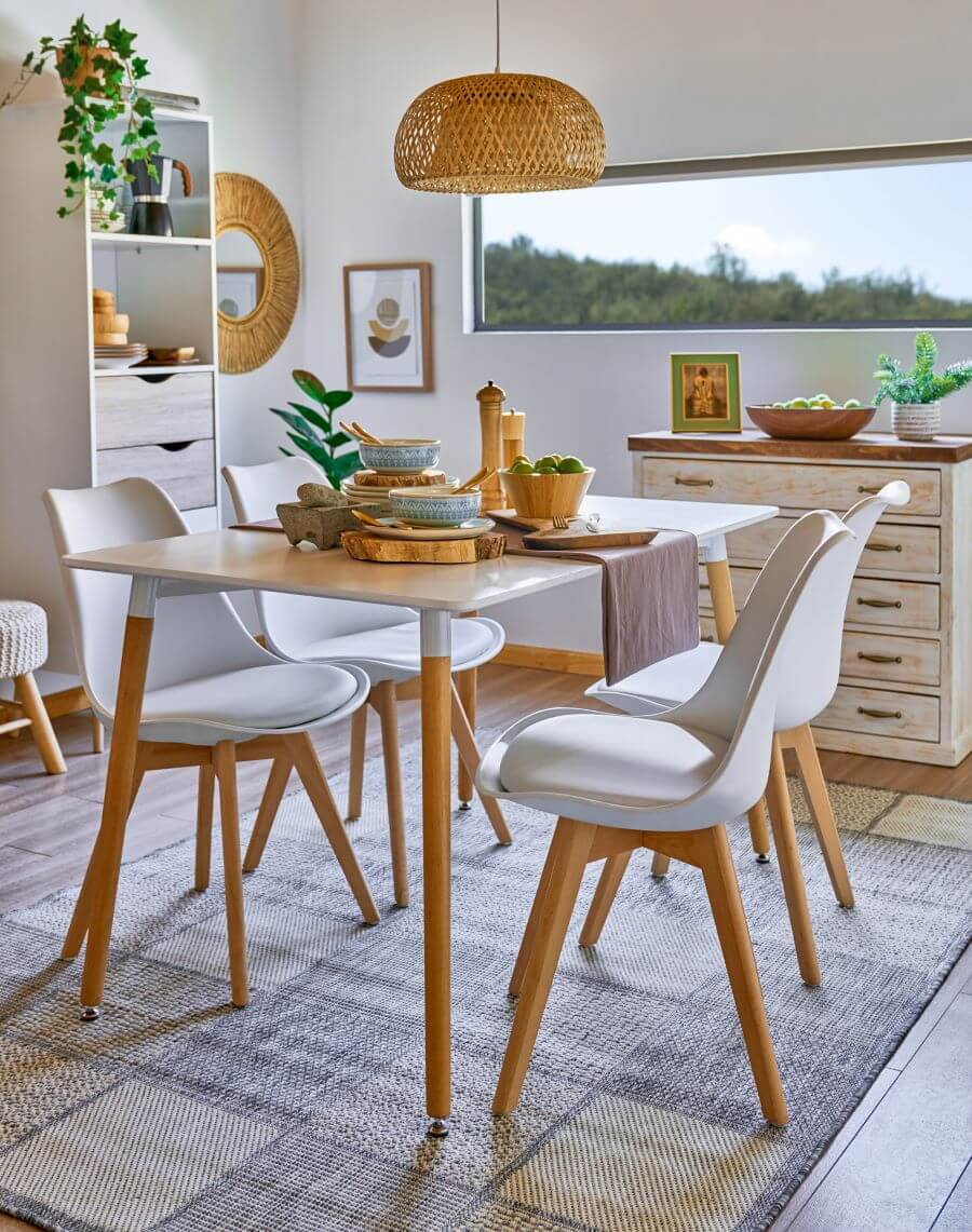 Comedor con mucha luz natural, muros blancos y ventana alargada en la parte superior. Comedor de base blanca con patas de madera, para 6 personas. Hay 4 sillas tipo "eames", con base blanca acolchada y patas de madera. Sobre la mesa hay vajilla de cerámica y una base de madera y bowls de madera. Sobre la mesa hay una lámpara colgante de mimbre. En el suelo hay una alfombra gris. Se ve un mueble con 5 cajones, en color beige desgastado y base de madera. Atrás del comedor hay una repisa con cajones y estantes, de color blanca, que tiene vajilla y una planta colgante.