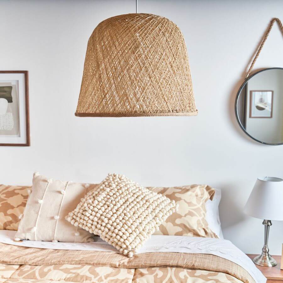 Dormitorio estilo boho. Gran lámpara de techo de fibras naturales en tonos café claro. Cama de dos plazas con cubrecama color beige y café claro, con dos cojines con textura. Muro blanco, con un cuadro y un espejo redondo con una cuerda para colgar. Junto a la cama hay un velador de madera y una lámpara de mesa de base metálica plateada y pantalla de tela blanca.