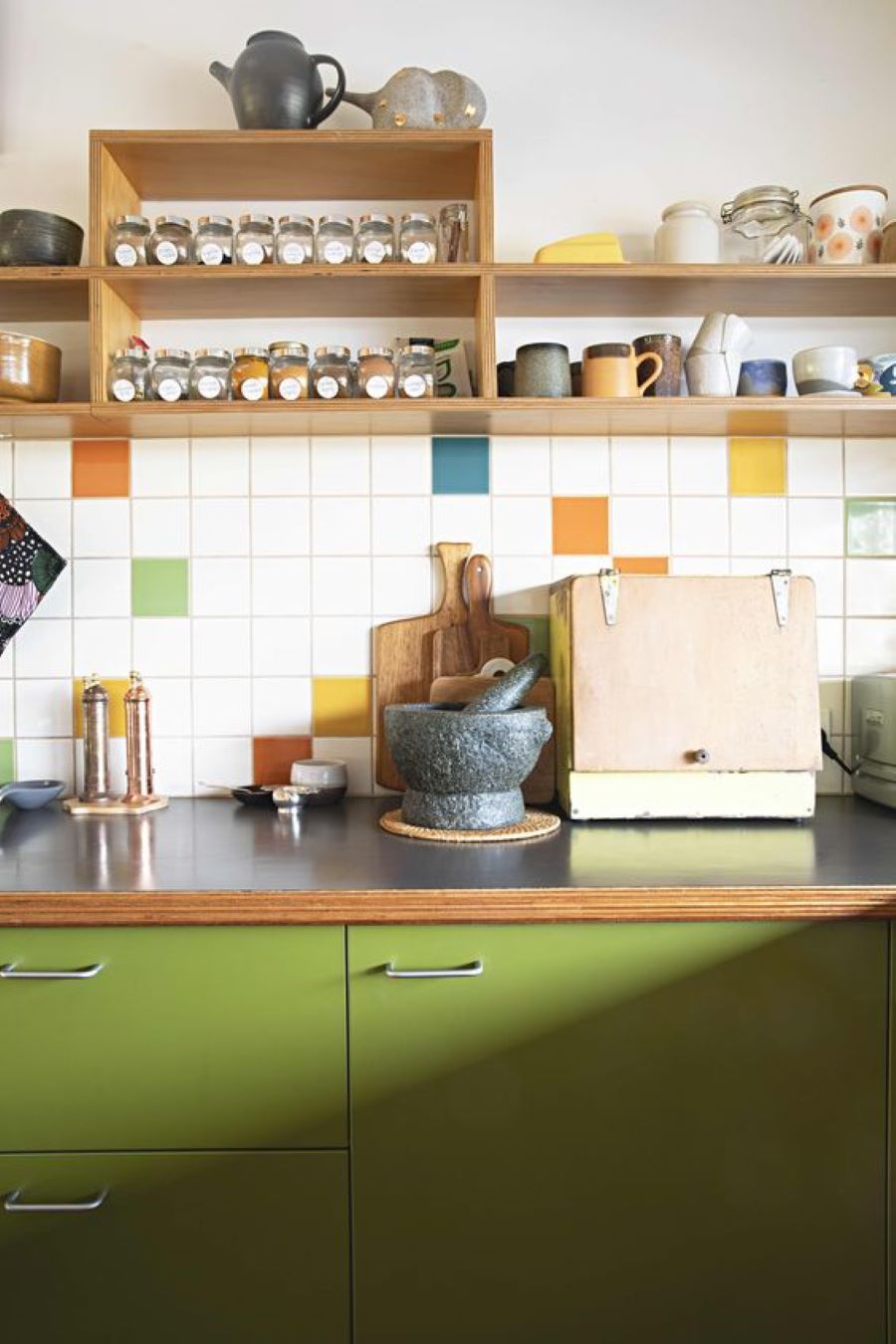 Cocina con muebles inferiores de color verde, mesón de madera. Muros con azulejos cuadrados blancos y de colores azul, naranjo, amarillo y verde. Repisa de madera, abierta, en la parte superior, con frascos de vidrio y tazones de cerámica. 