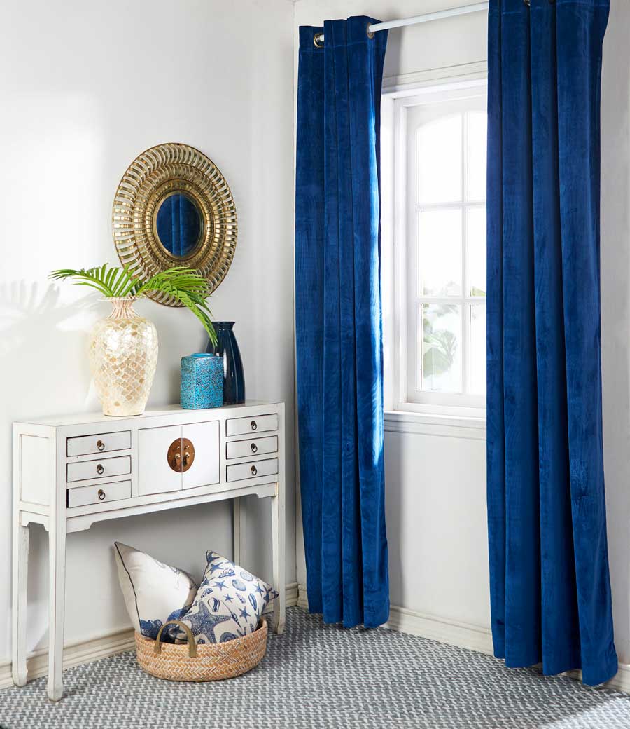 Rincón del hogar con una ventana con cortina velvet azul, que ayuda a ahorrar energía. También hay un arrimo blanco y sobre ella un espejo redondo con marco dorado.