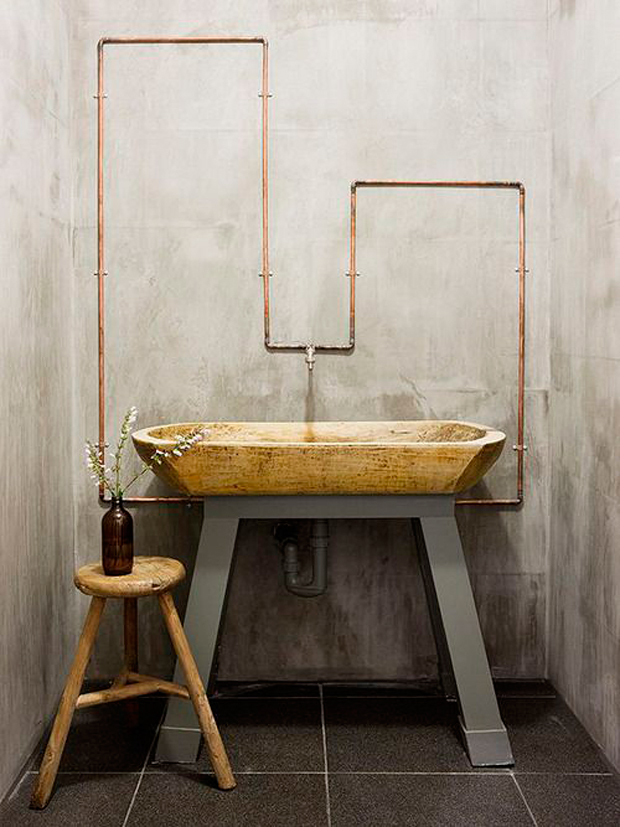 Baño con un lavamanos, piso de madera a su costado y sobre é, una cañería de cobre que forma un diseño para darle un nuevo look a la pared de concreto.
